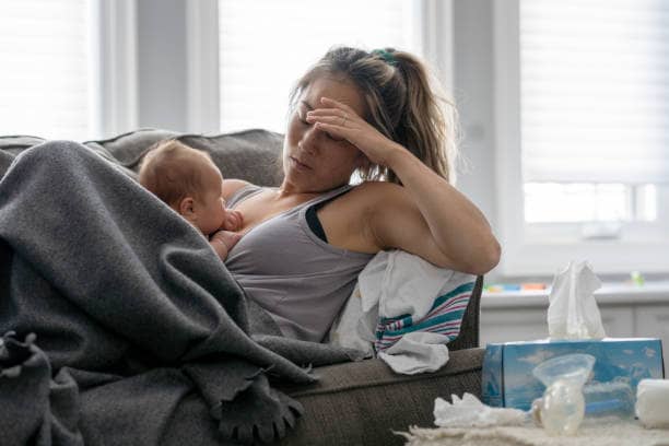 A tired mother breastfeeding her baby on a couch while resting under a blanket, with tissues and baby supplies nearby, capturing the exhaustion and emotional weight of postpartum motherhood.