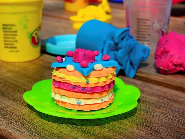 Colorful layered Play Doh cake on a green plate, decorated with small Play Doh dots, with Play Doh containers and tools blurred in the background.