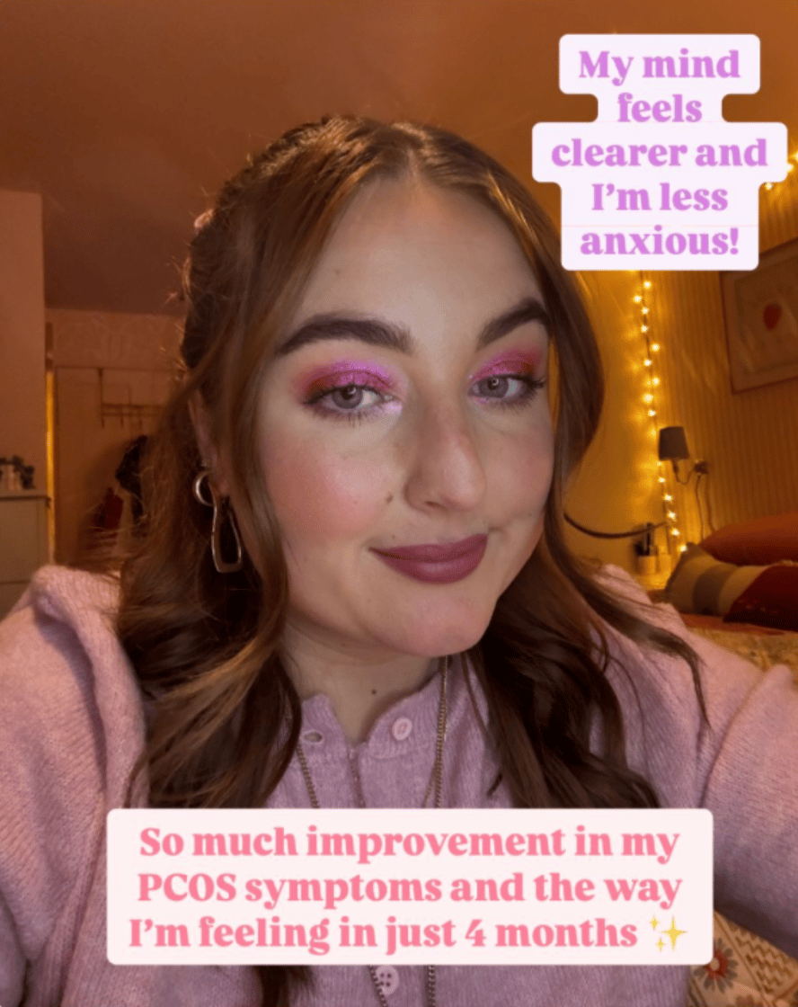A woman with long brown hair is smiling at the camera, wearing pink eyeshadow and lipstick. She is dressed in a light pink sweater. The room has warm lighting with string lights in the background. Text on the image reads: "My mind feels clearer and I’m less anxious!" and "So much improvement in my PCOS symptoms and the way I’m feeling in just 4 months."