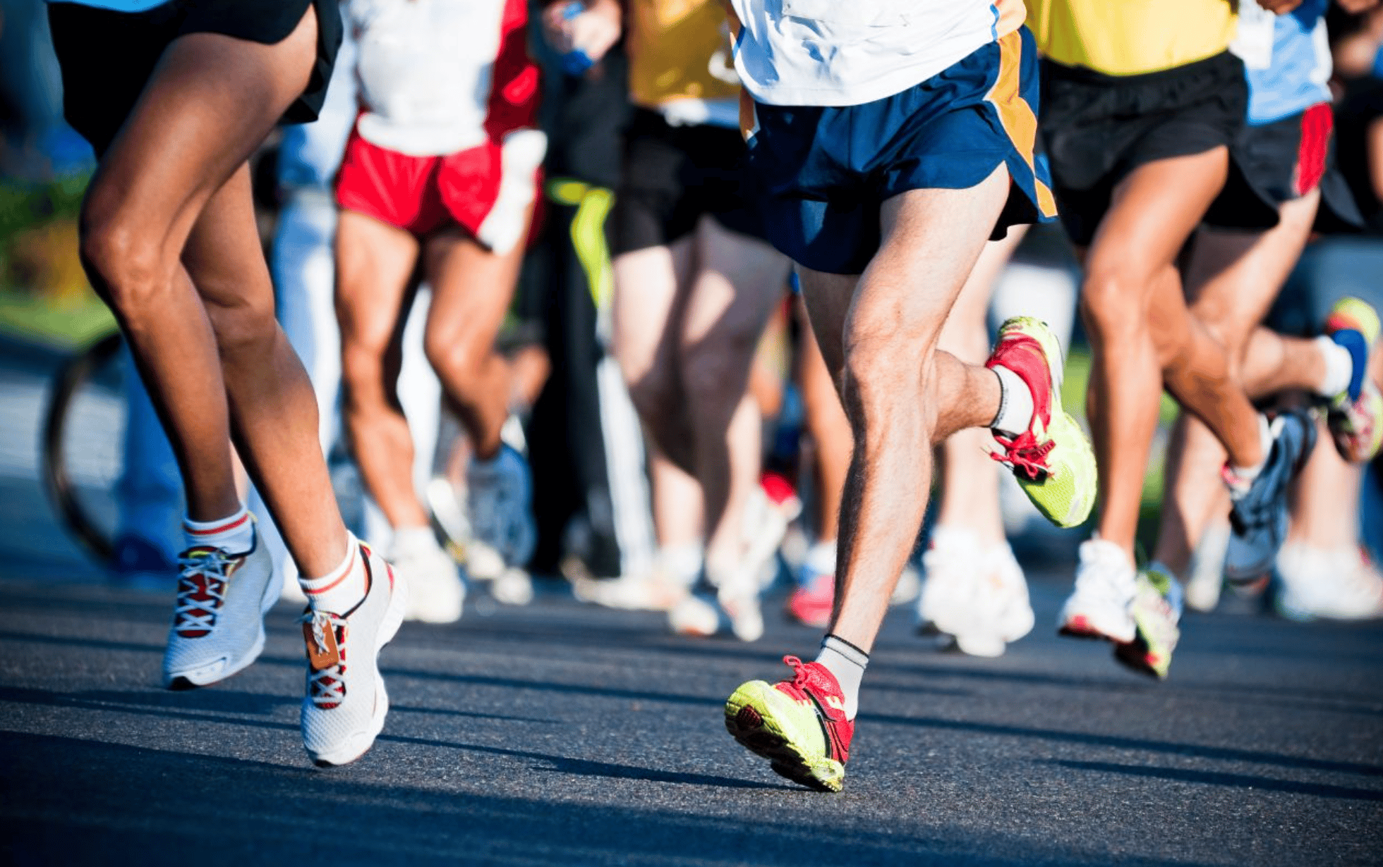 A group of runners in motion, captured from the waist down, showcasing various colorful running shoes. They are running on a paved road, indicating a race or marathon event. The focus is on their legs and footwear against a blurred background.