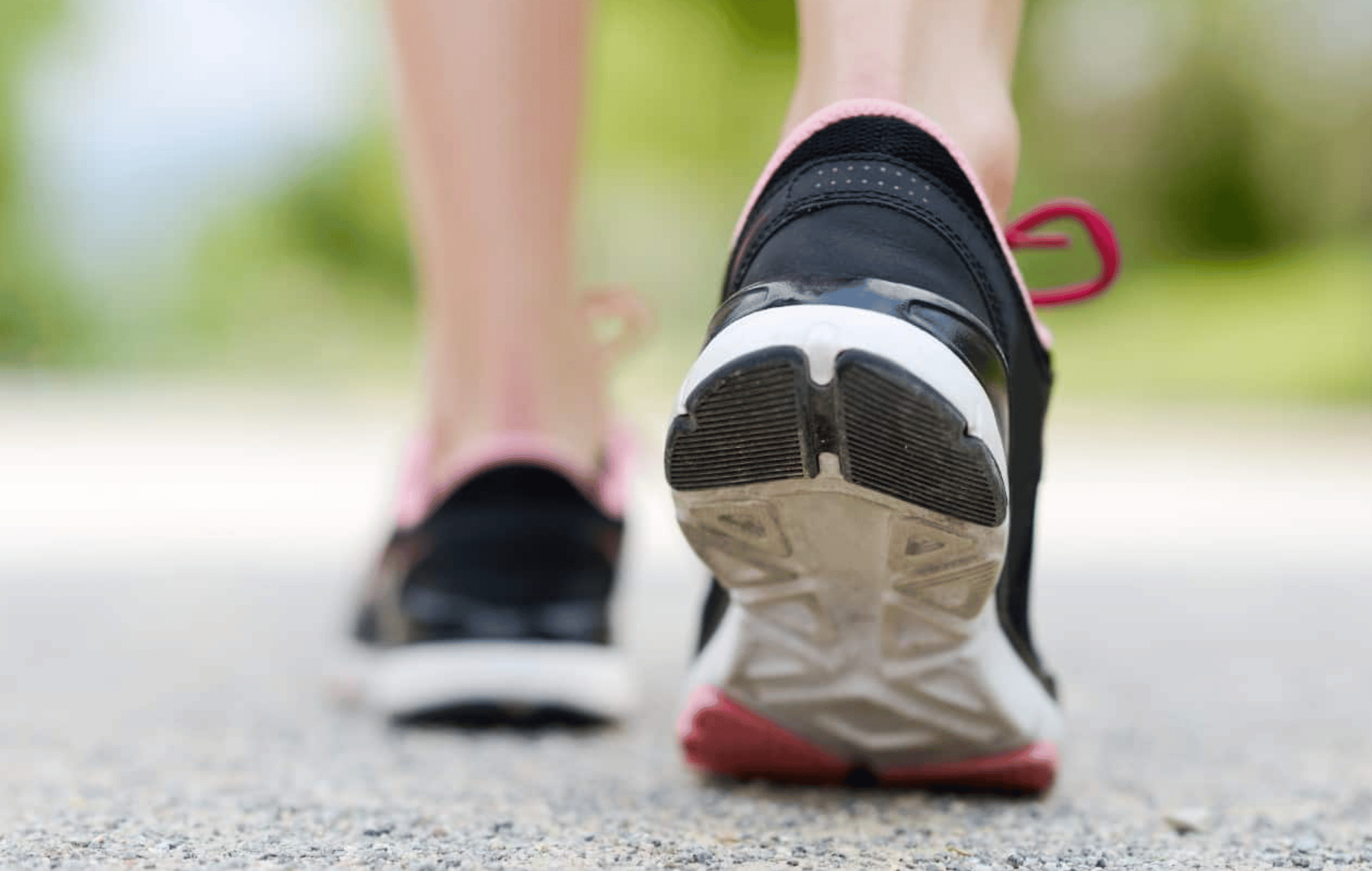 Close-up of a person walking on a path wearing black running shoes with pink accents. The focus is on the sole of the shoe, showing a textured pattern. The background is blurred with green foliage.