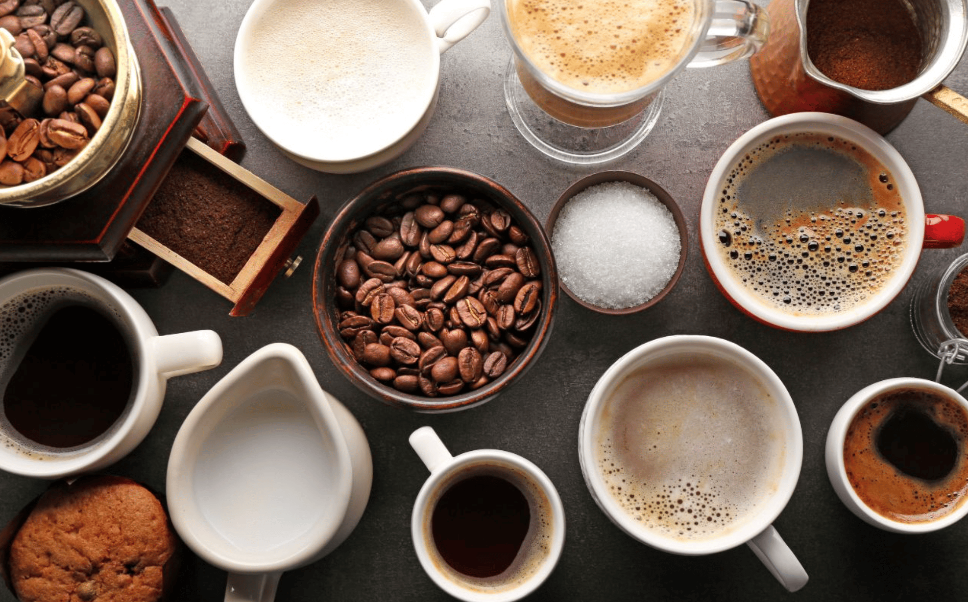 An assortment of coffee items on a table, including various cups of coffee with different levels of crema and froth. There's a bowl of whole coffee beans, a small cup of sugar, a milk jug, and freshly ground coffee displayed next to a grinder. A cookie is also visible, adding a touch of sweetness.