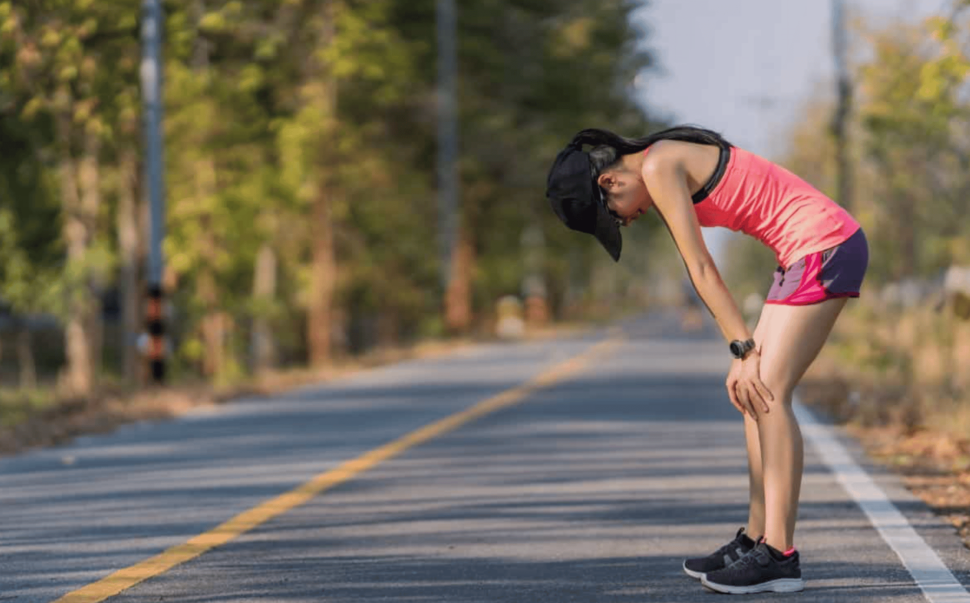 A woman wearing a pink tank top, purple shorts, and a black cap is bent over, resting her hands on her knees, appearing tired or taking a break while running on a road lined with trees. The road is empty, with a clear yellow line running down the middle, and the background features blurred green foliage.