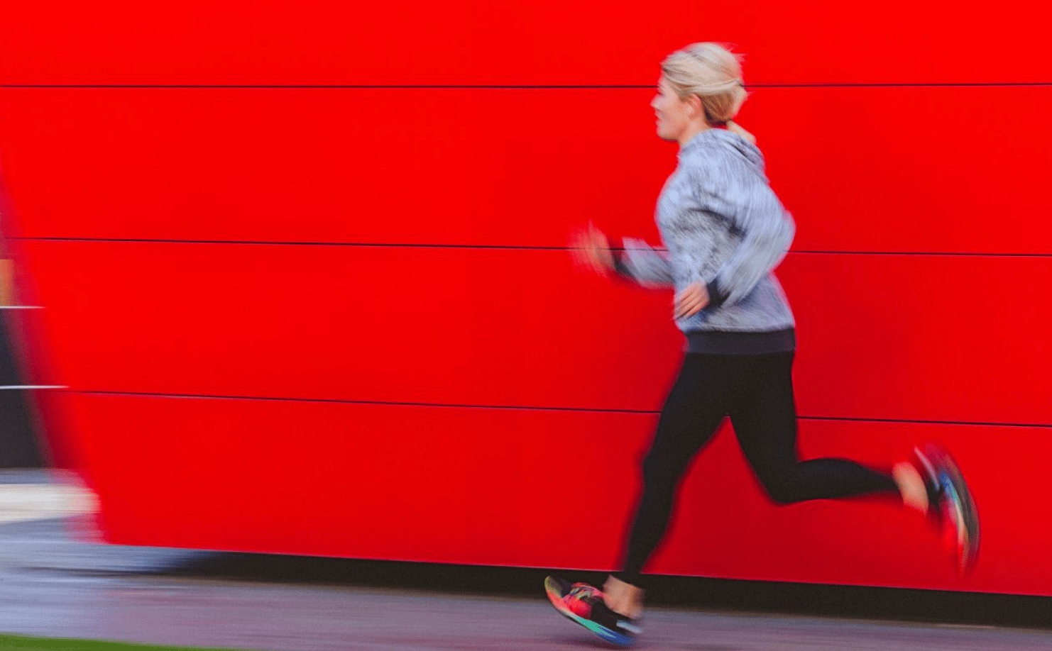 A person in athletic clothing is running past a bright red wall. They wear a gray hoodie, black leggings, and colorful running shoes. The motion blur gives a sense of speed and movement.