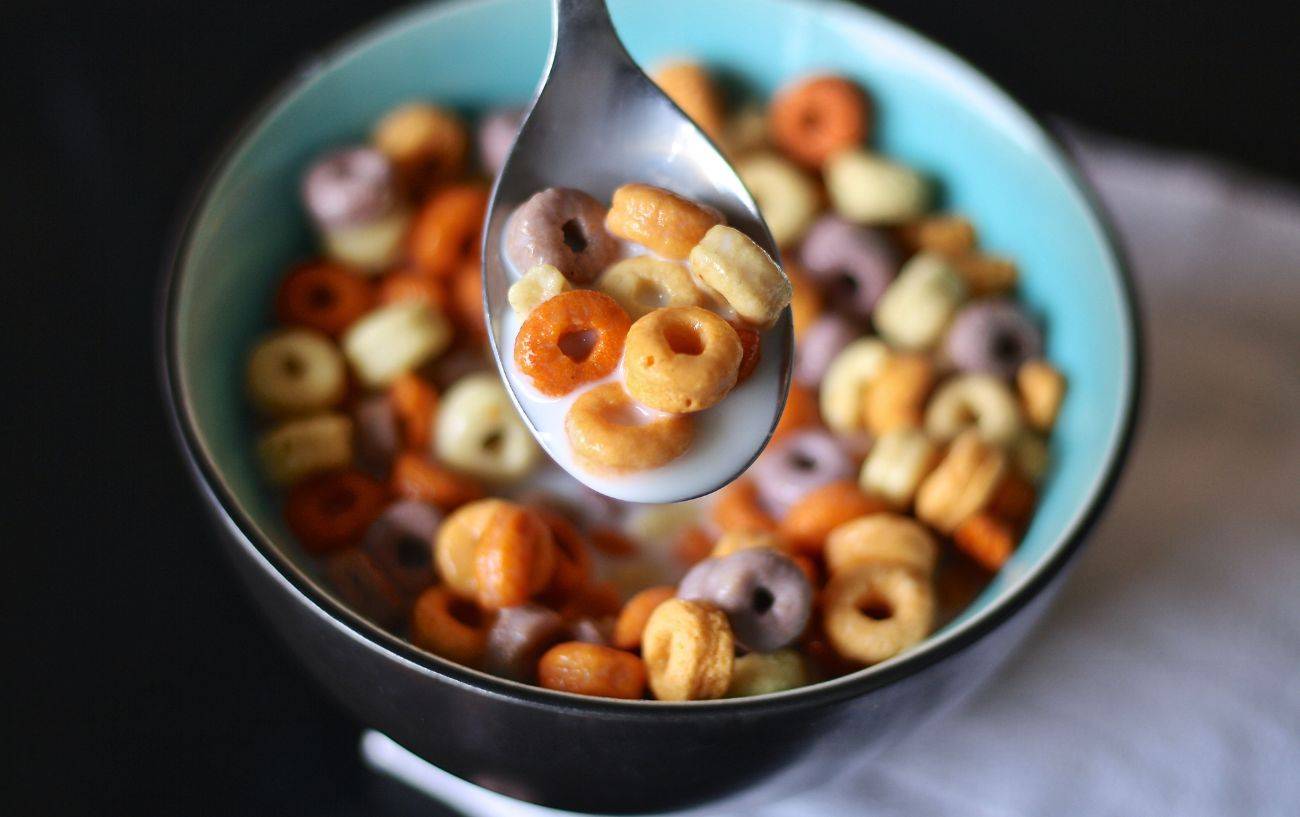 A close-up image of a bowl filled with colorful, round cereal pieces in milk. A spoon is being lifted from the bowl, containing several cereal loops in shades of orange, yellow, and purple. The bowl is blue on the inside and is set against a dark background with a hint of a napkin underneath.