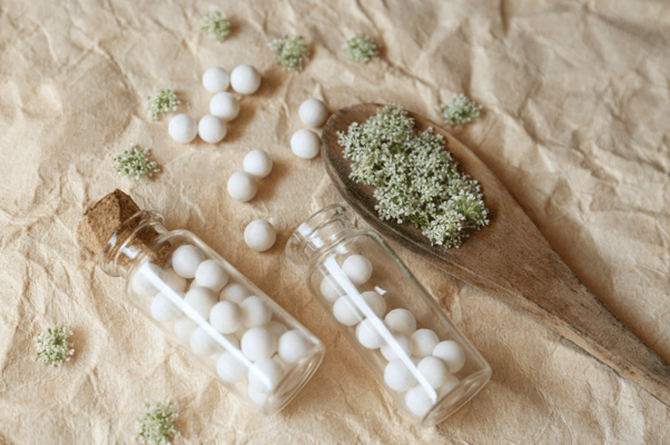 Glass bottles with white homeopathic remedies (globules) and small flowers on a textured fabric.