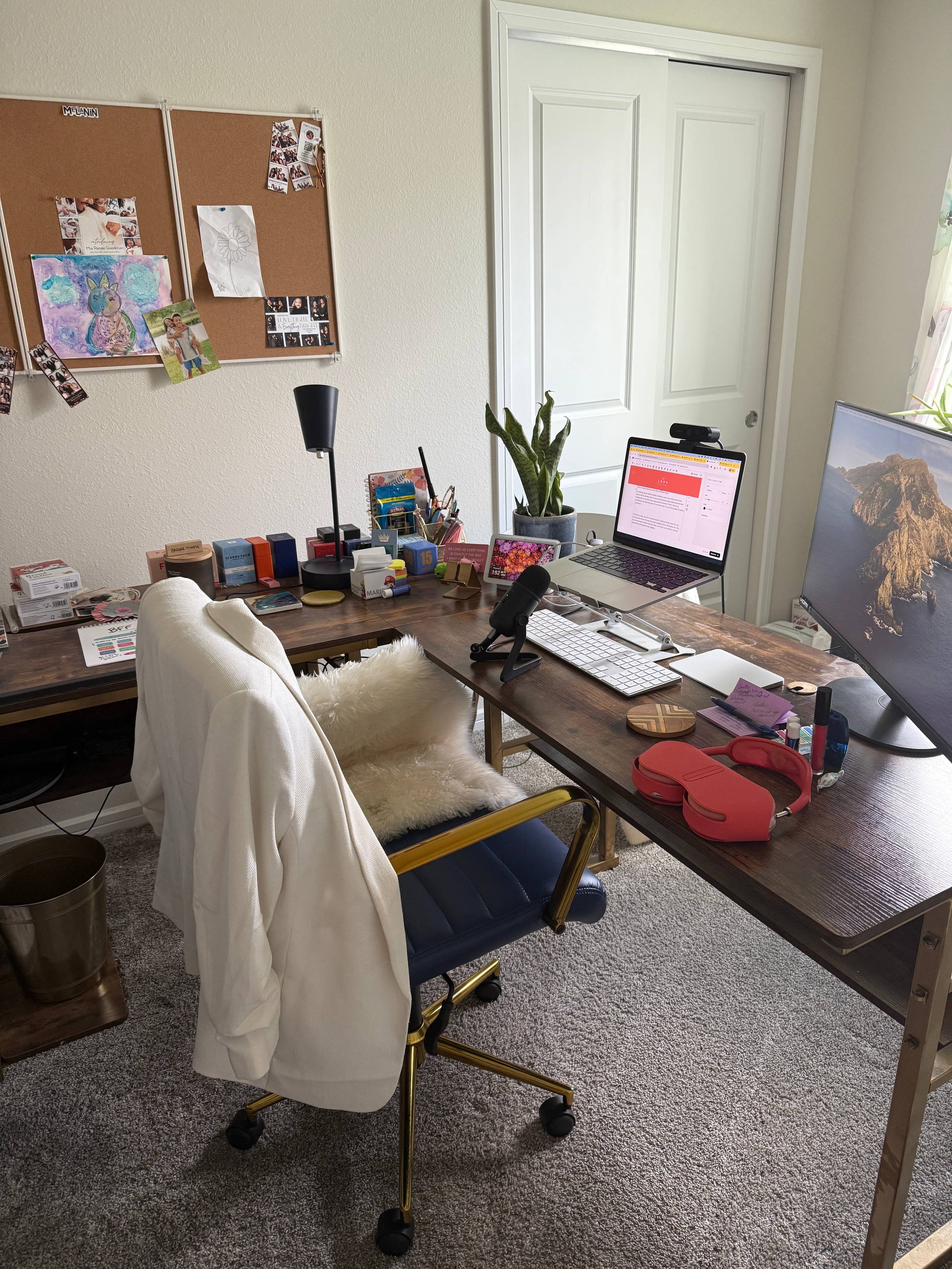 This image shows a cozy, well-organized home office.  A white blazer hangs neatly over the back of a navy-blue, gold-framed desk chair with a fluffy white seat cushion. The desk itself is dark wood with gold legs, and it holds two computer screens — a lap