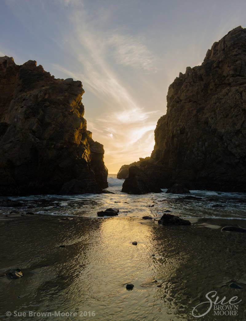 A photograph of sunset at Big Sur, California with the waves receding toward a gap in the rocks