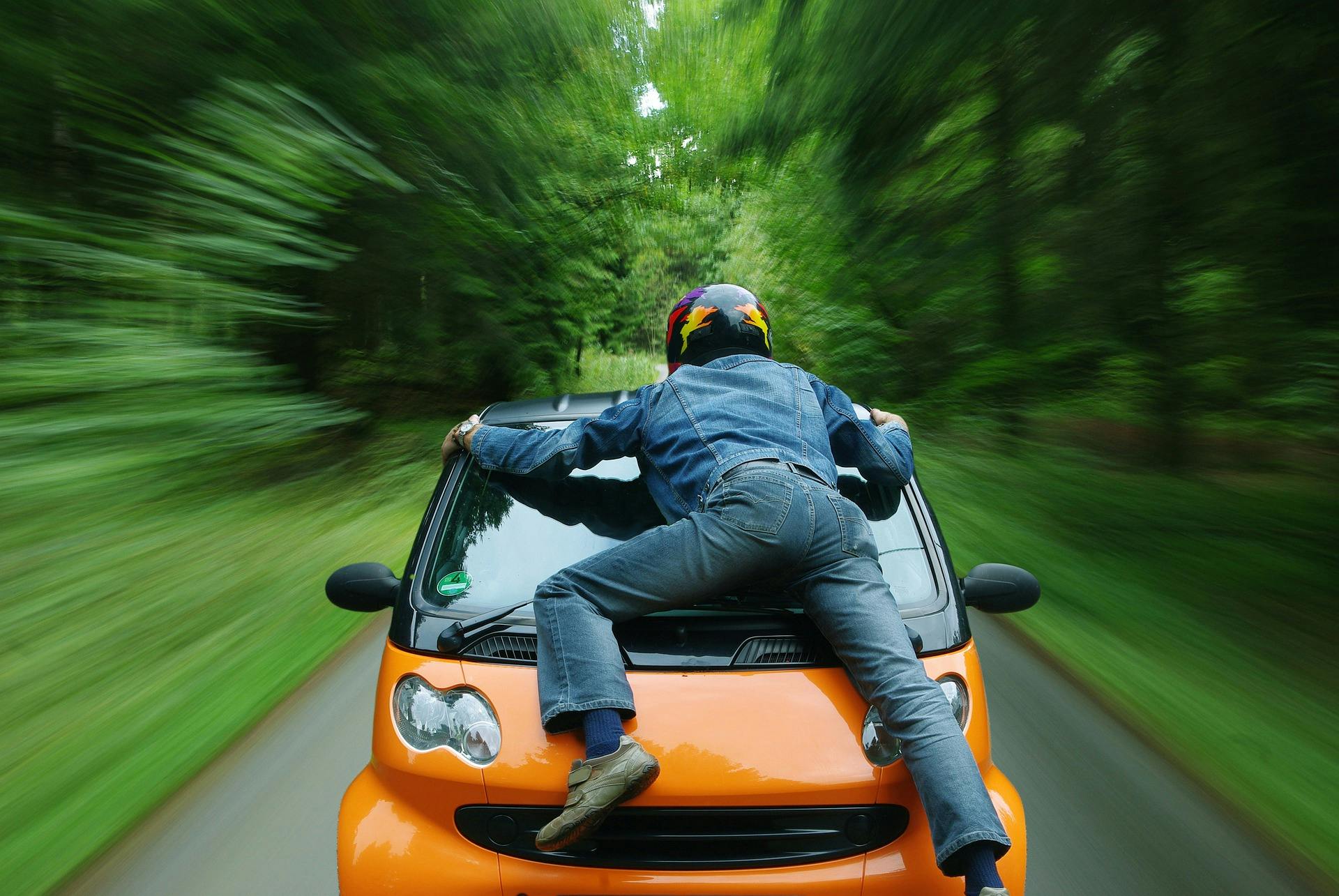 Man on a car's hood, holding on as it speeds through a forest 