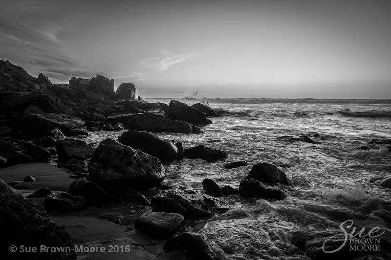 A photograph of sunset at Big Sur, California with the waves crashing onto a rocky shore in monochrome