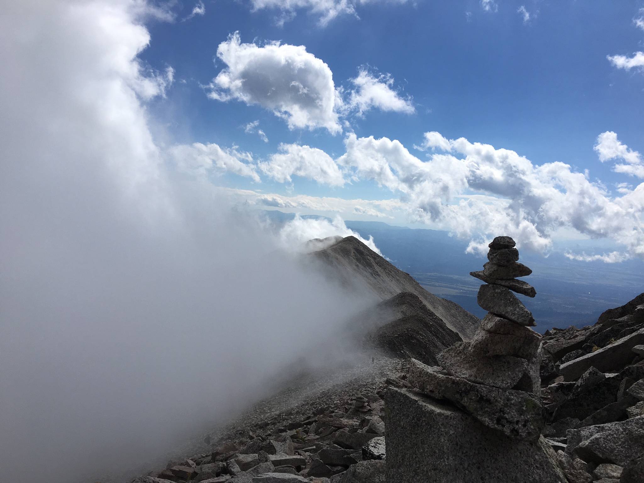 A grey mist creeps up the side of a rocky mountain ridge that extends into the blue sky speckled with bright white clouds in the background. A tower of flat rocks balances in the foreground.
