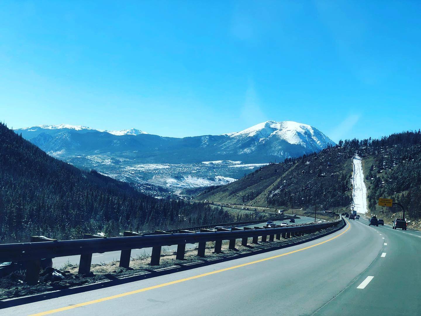 A smooth highway in the foreground leads downhill toward a panorama of tall, snowy mountain peaks, above deep green pine forests with a snowy emergency ramp for trucks in view and the clearing of a town at the foot of the mountains in the distance.