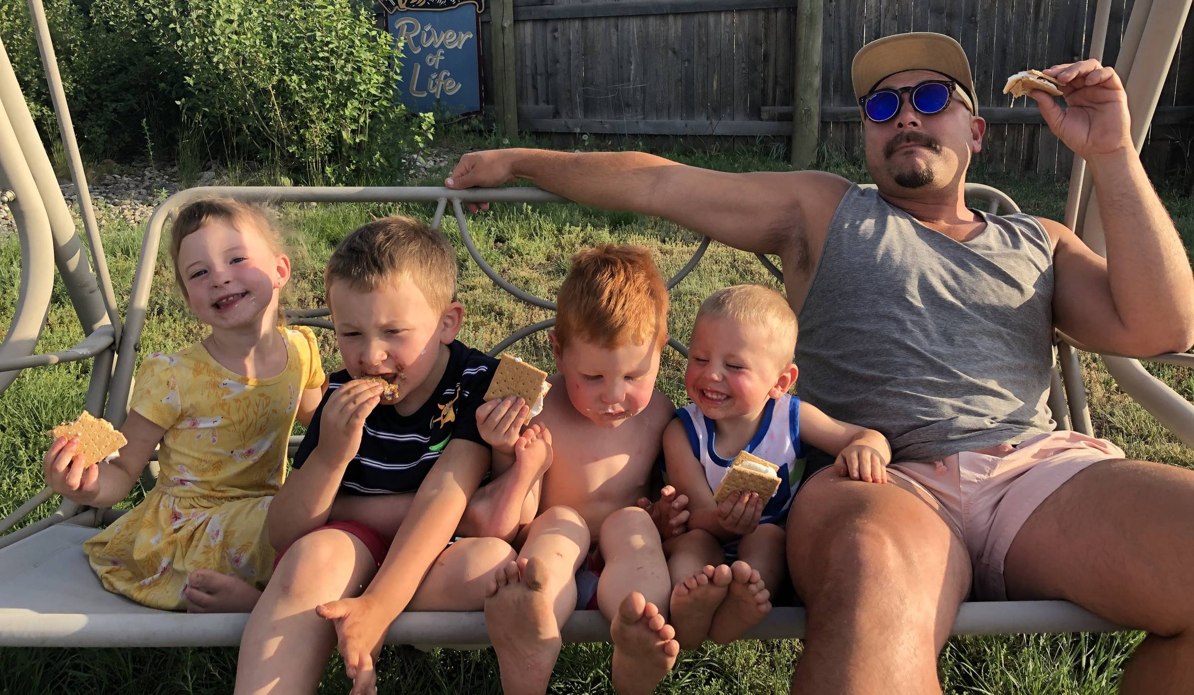 Four children crowd onto a swing chair next to an adult with a goatee in sunglasses and a cap, and everyone has s'mores in their hands and chocolate and marshmallow on their faces.