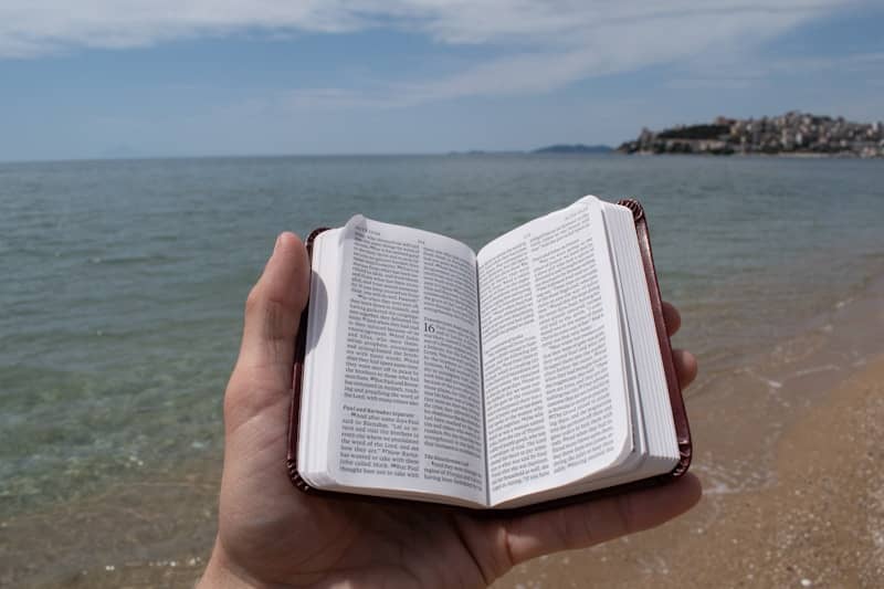 A hand holds an open bible at the beach.