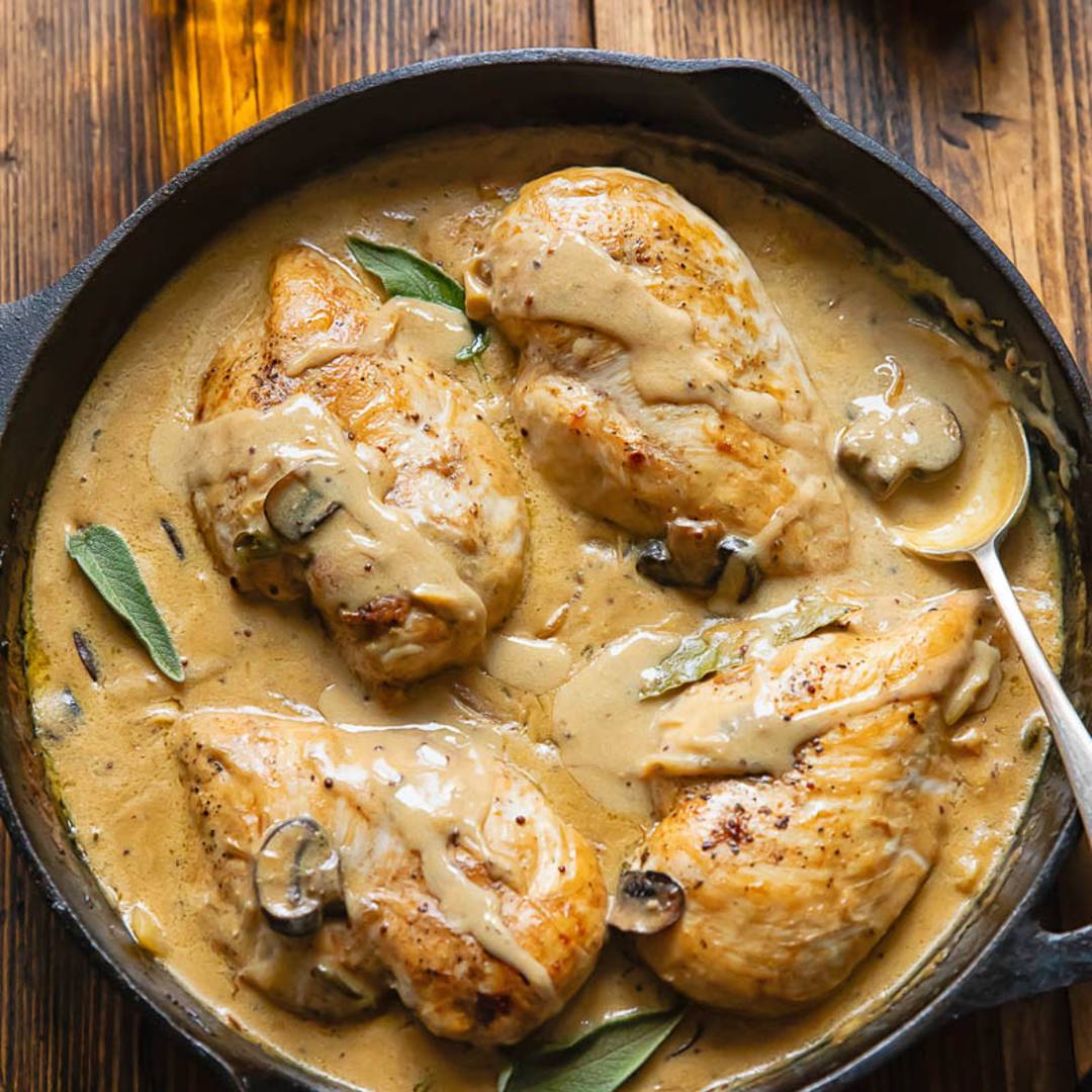 Cast iron skillet on a wooden background, with four chicken breasts in a creamy sauce.