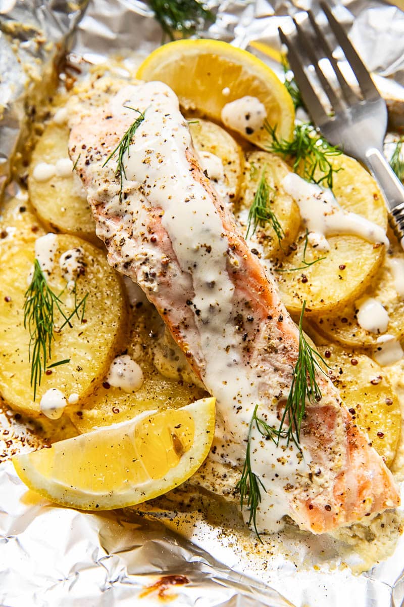 a piece of salmon with sauce and dill on top of sliced potatoes in aluminum foil