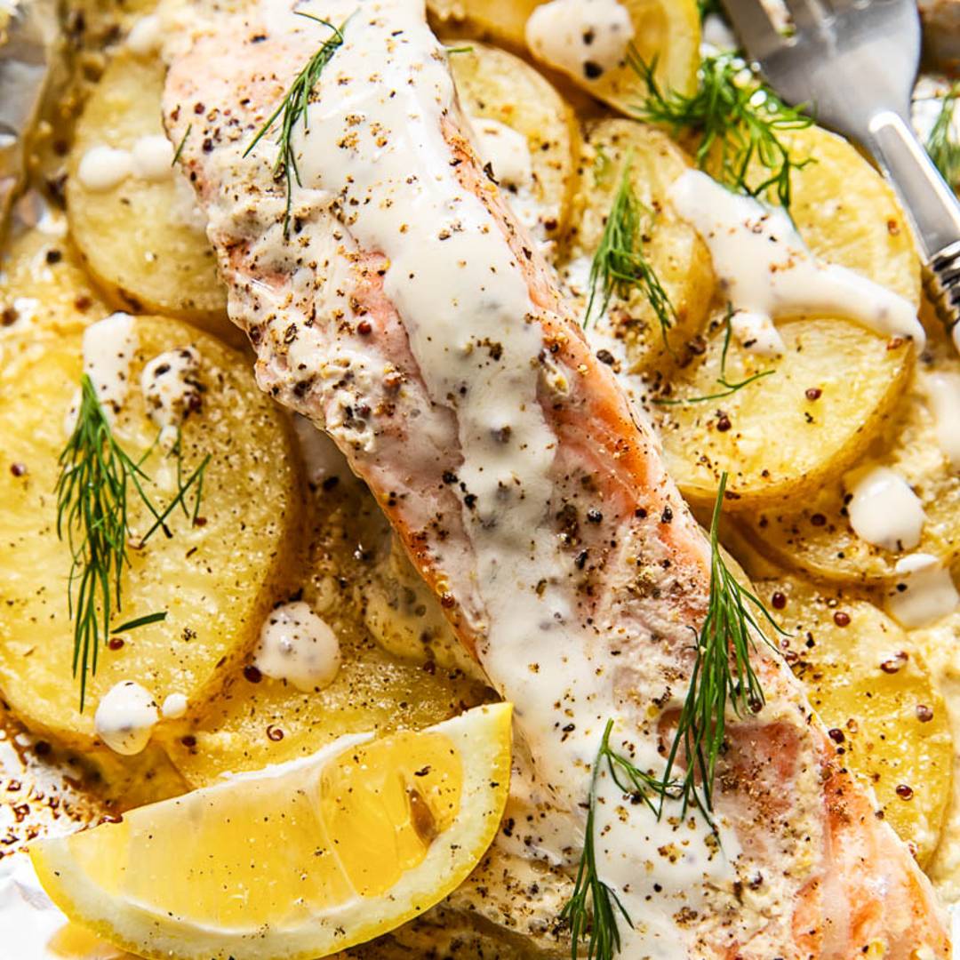 a piece of salmon with sauce and dill on top of sliced potatoes in aluminum foil