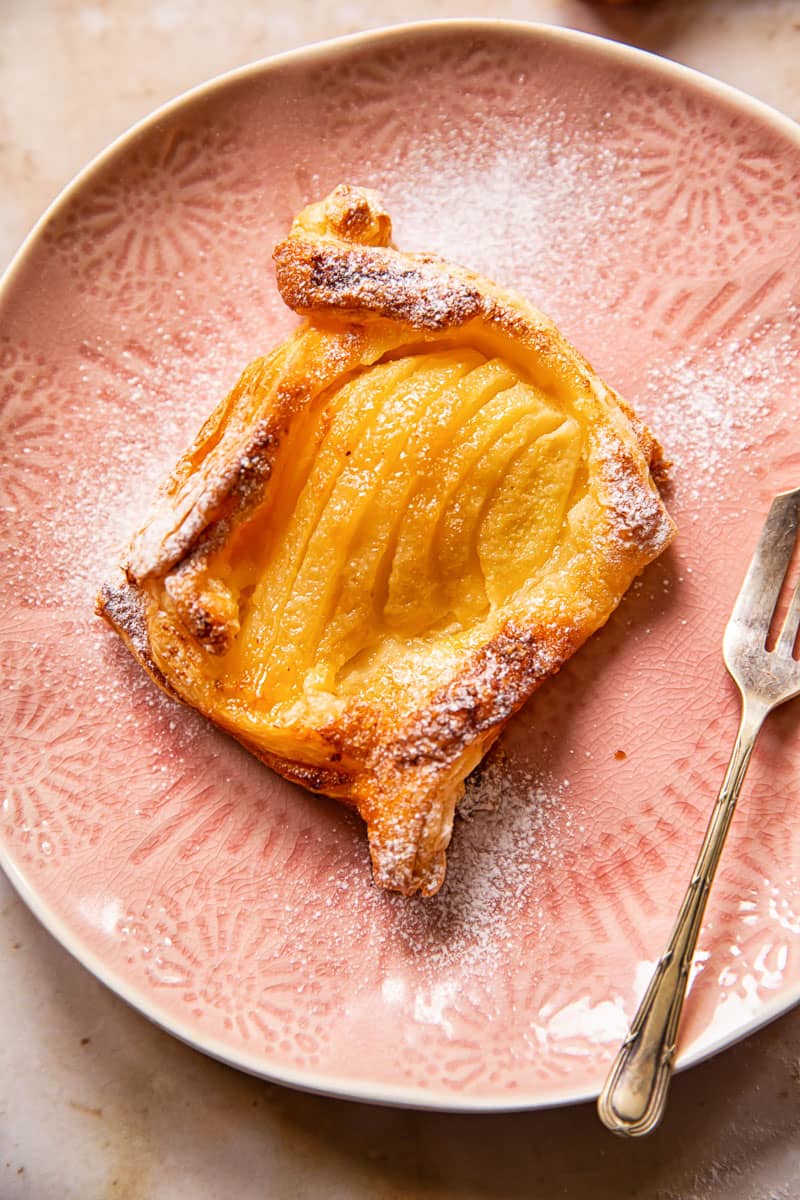 a pear tart, dusted with icing sugar, on a pink plate with a small dessert fork