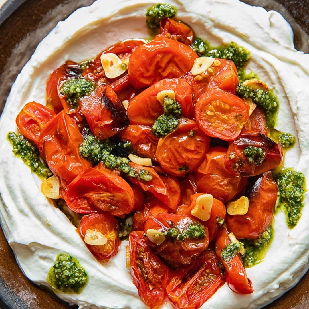 a white dip on a dark plate, topped with halved cherry tomatoes and basil pesto