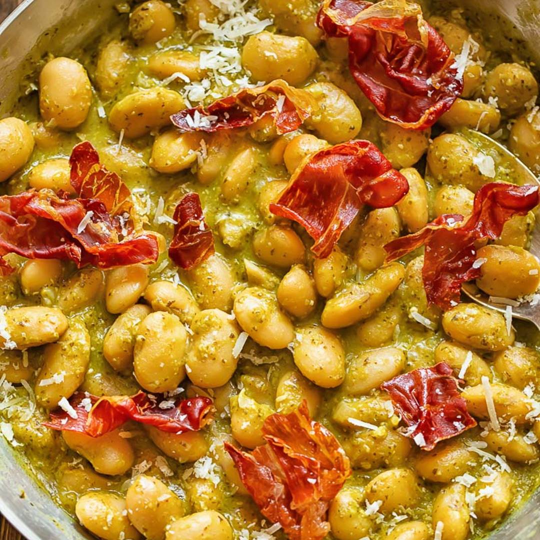 pan with beans in a green sauce with pieces of prosciutto on top