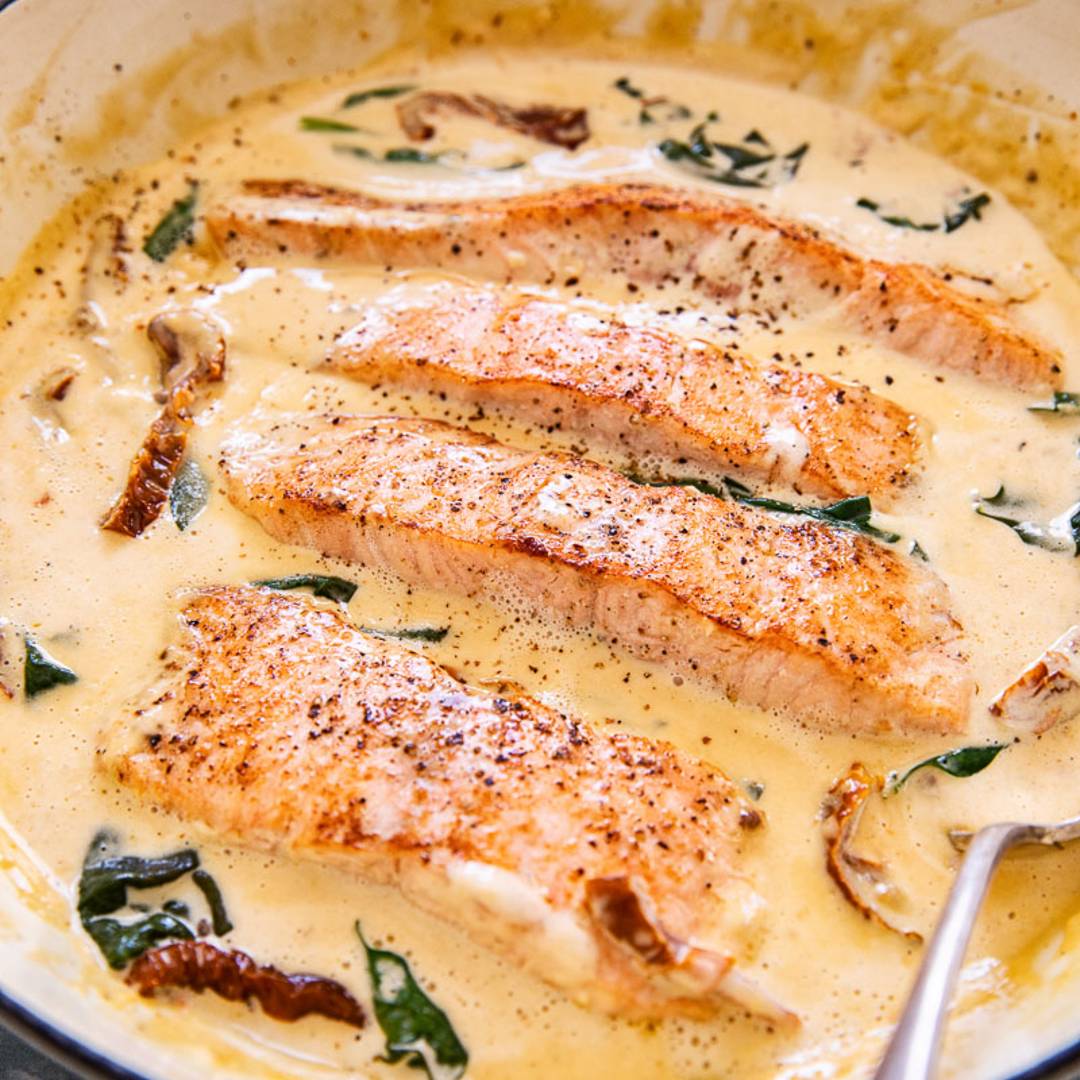 a large pan with four salmon pieces in a creamy sauce with greens, sun-dried tomatoes and a serving spoon