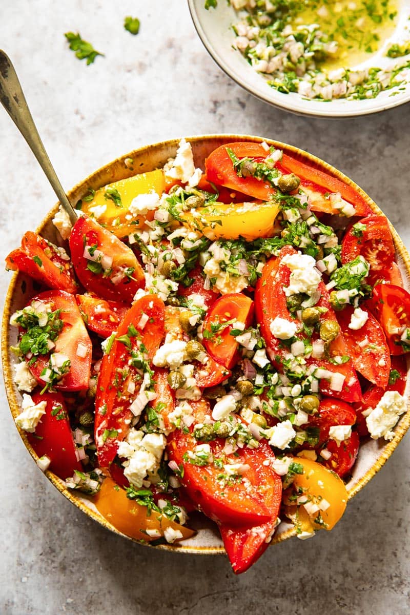 top down view of a bowl of tomato salad topped with herbs and crumbly cheese, with a serving utensil