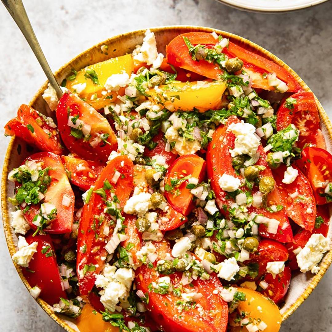 top down view of a bowl of tomato salad topped with herbs and crumbly cheese, with a serving utensil