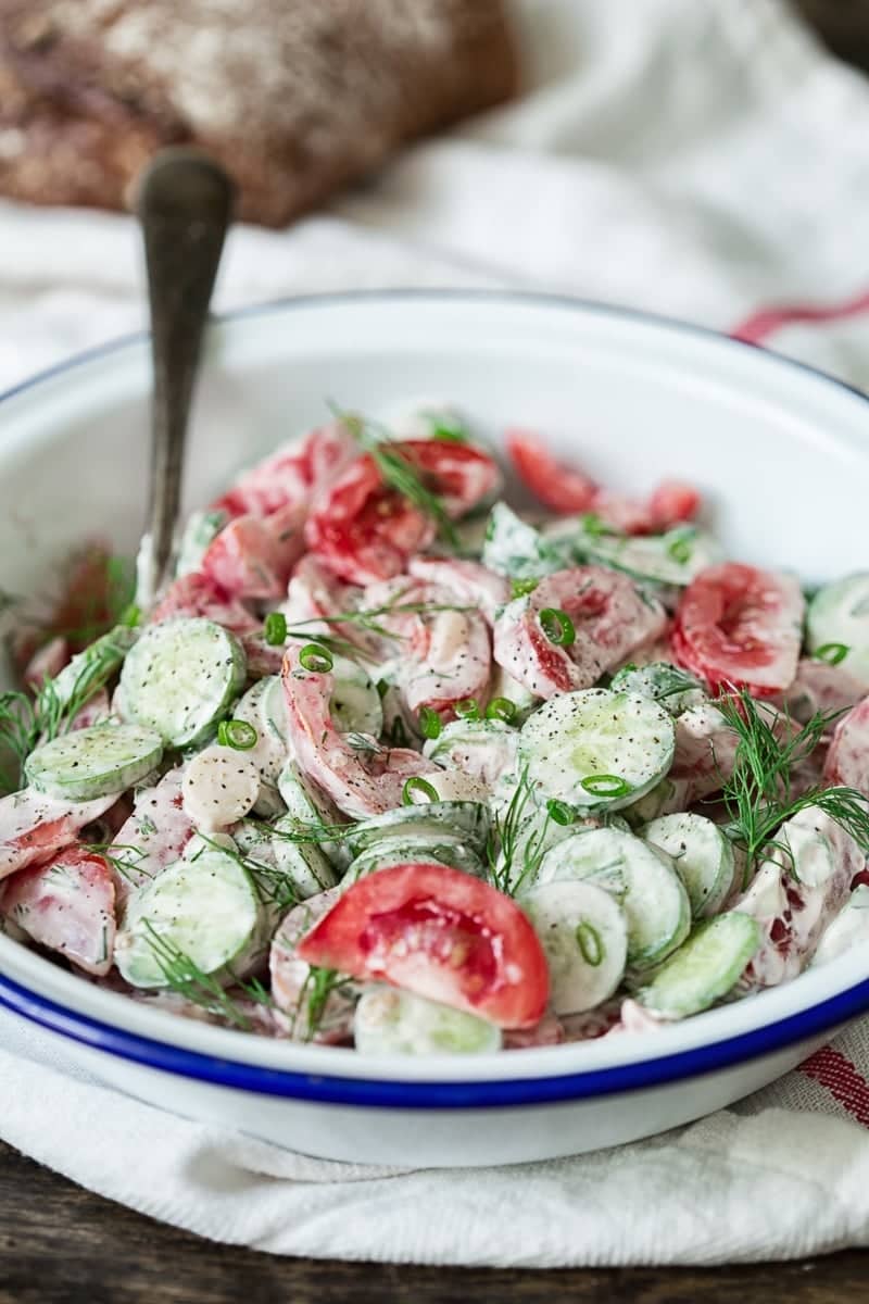 Bowl of salad with cucumbers and tomato in a white dressing and topped with dill.