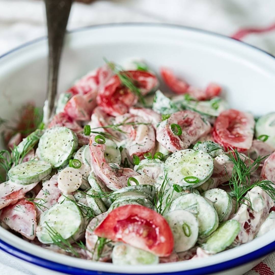 Bowl of salad with cucumbers and tomato in a white dressing and topped with dill.