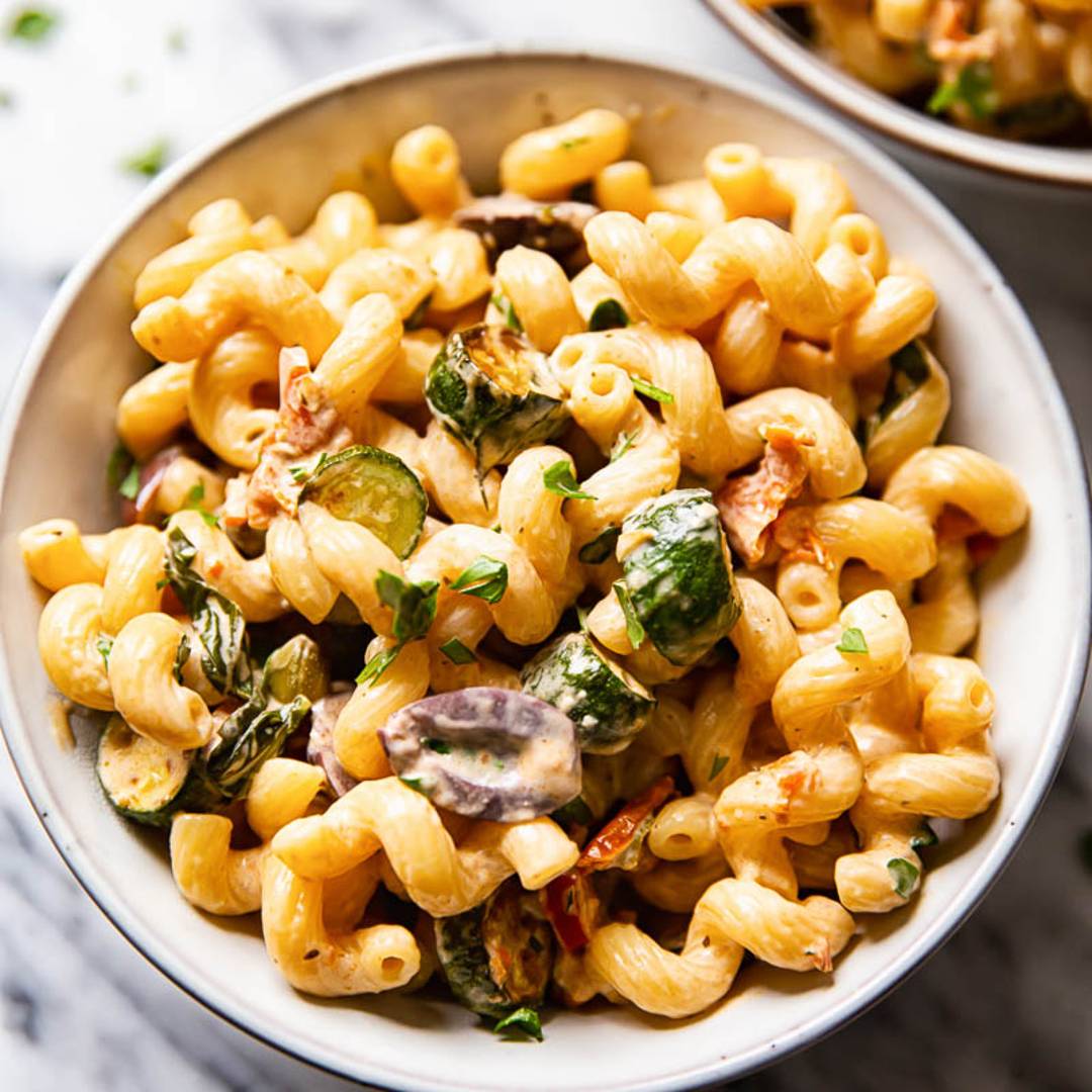 a bowl of curly pasta with zucchini and red olives on a light marble background