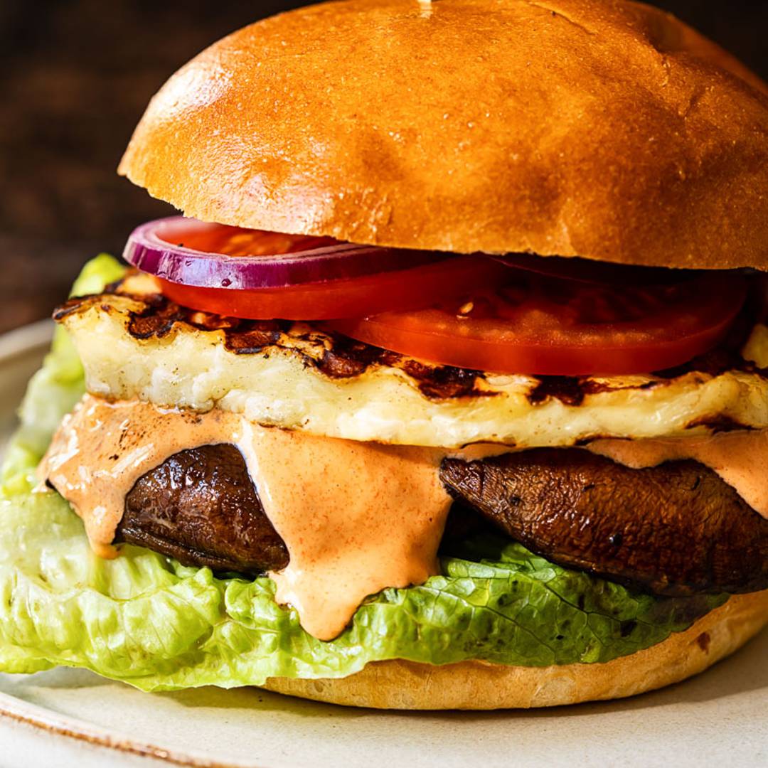 burger with brioche buns with layers of lettuce, mushroom, halloumi, tomato and onion, with a dripping orange sauce