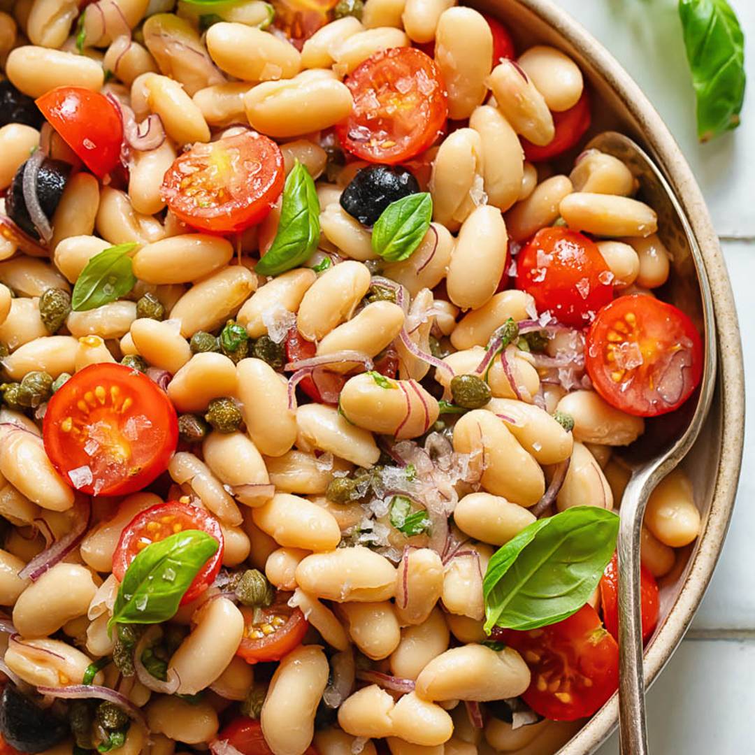bowl with white beans, tomatoes and olives topped with basil, with a serving spoon