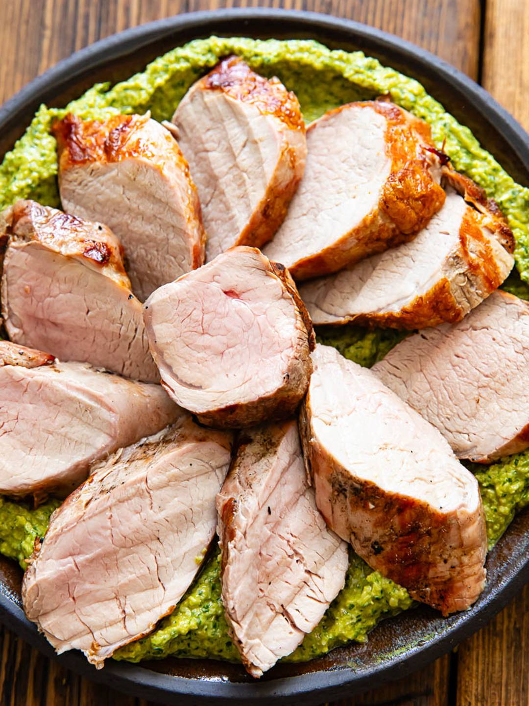 Slices of pork tenderloin on a bed of green sauce, on a plate with a dark wood background.