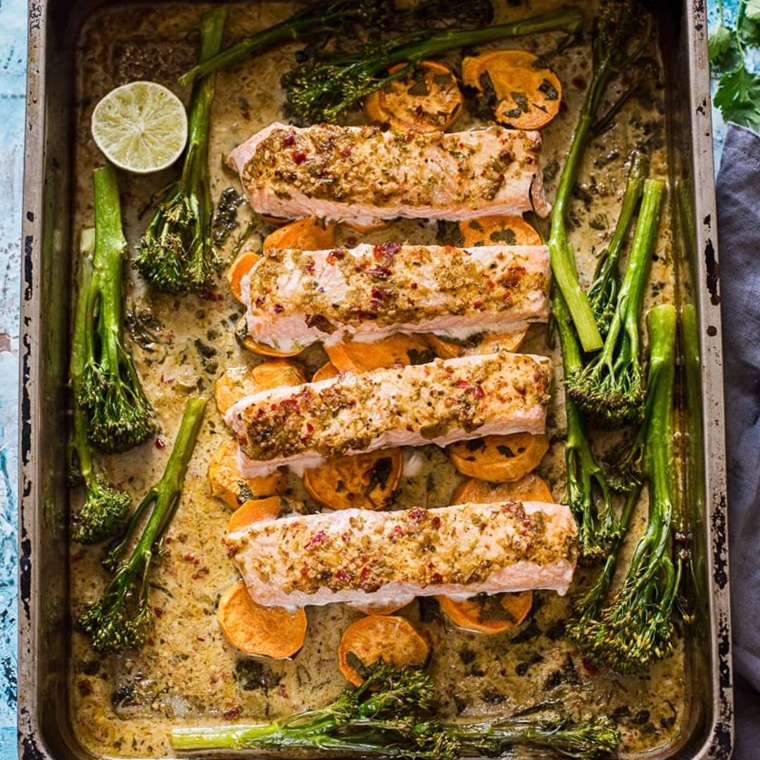 a baking pan with four salmon pieces layered on sweet potato slices with broccoli and a golden sauce