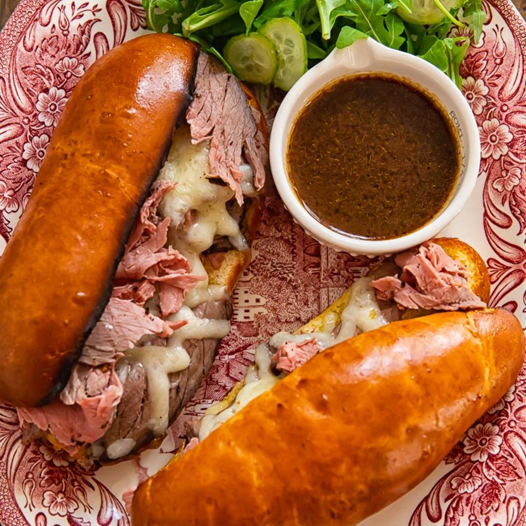two sandwiches with sliced roast beef and cheese in golden buns on a platter with gravy and a green salad
