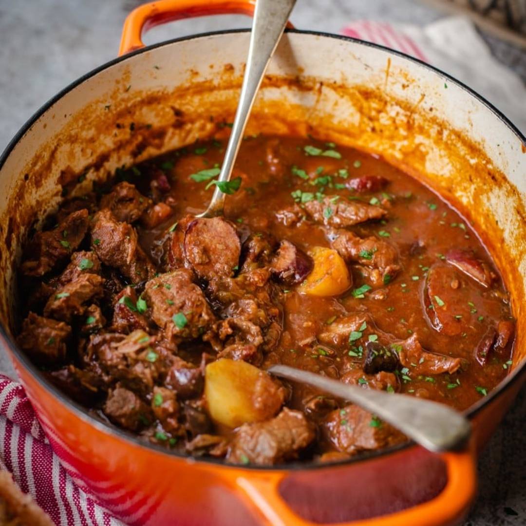 beef stew in an orange cast iron pot with two serving spoons