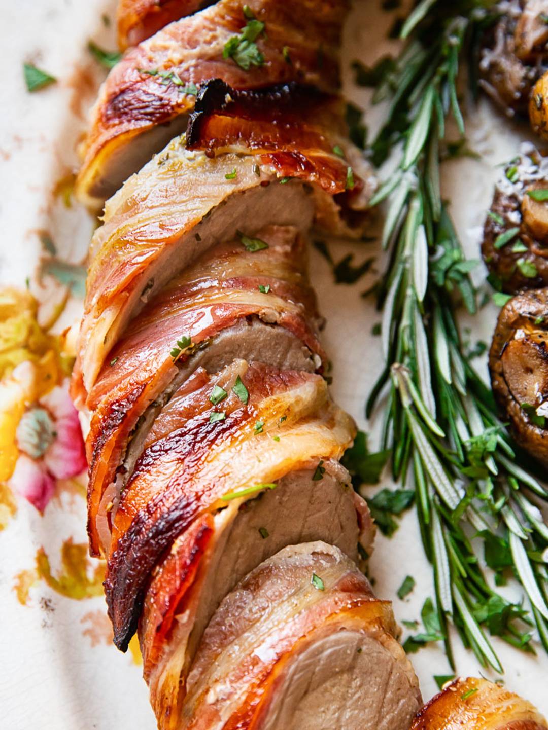 sliced pork tenderloin wrapped in crispy bacon, on a platter with a rosemary sprig.