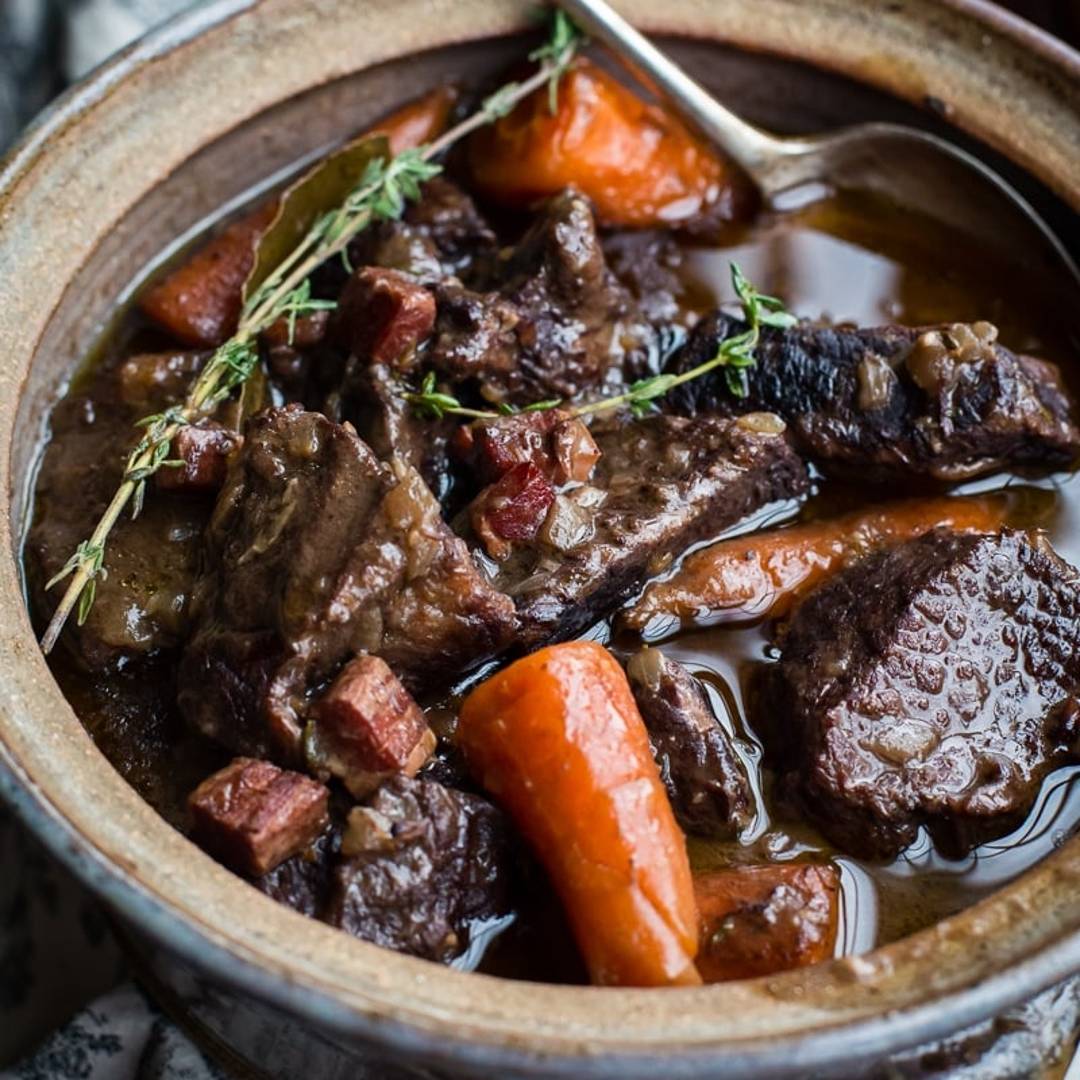 Beef Bourguignon in a clay pot with a serving spoon and sprig of thyme