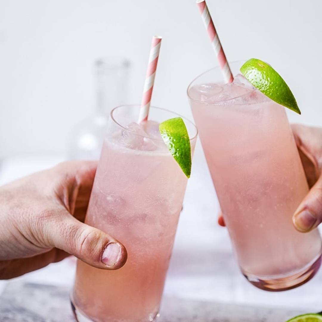 hands holding two glasses of a fizzy pink drink, with straws and lime slices as garnish