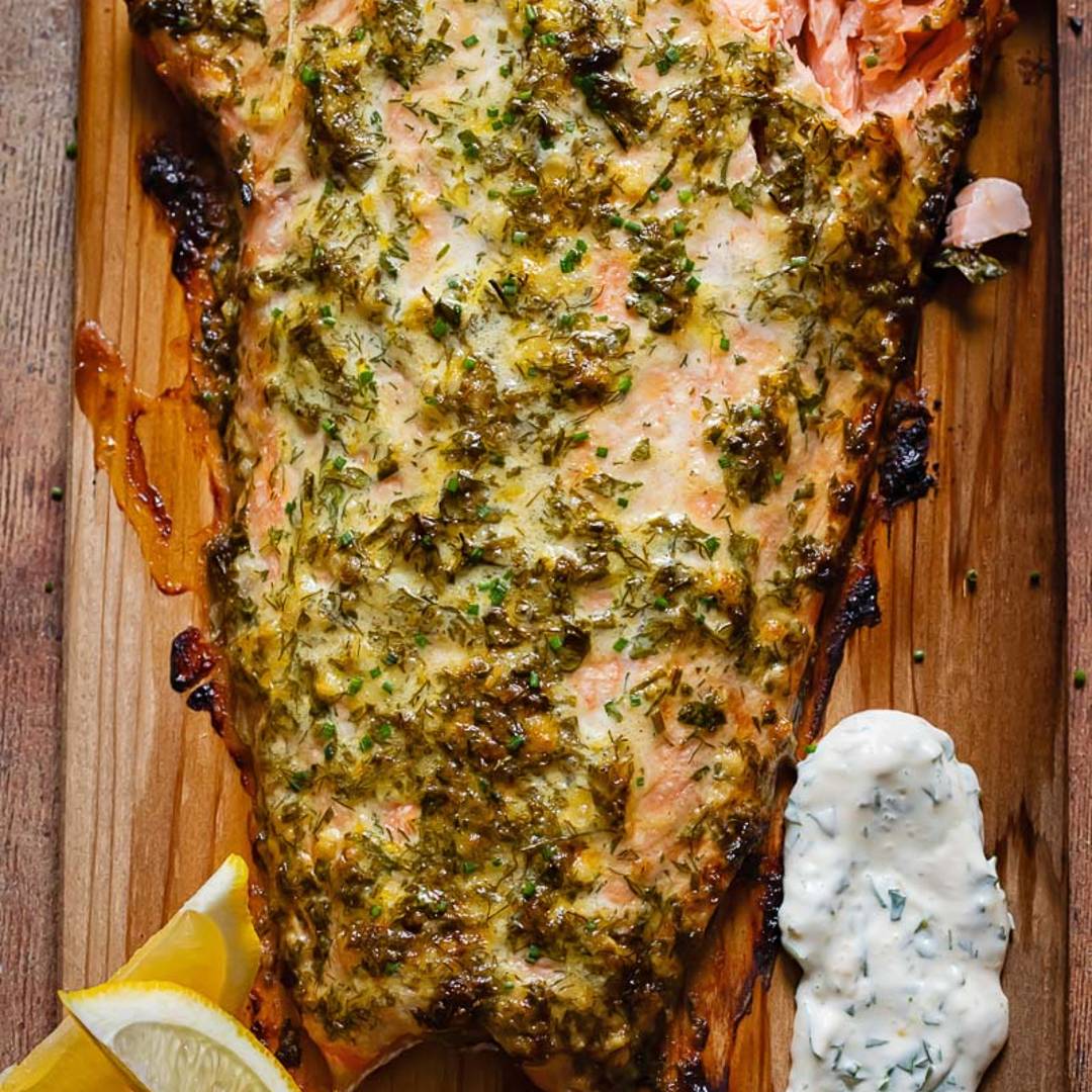 large slab of salmon on a wooden board, topped with greed spread and alongside lemon and yogurt