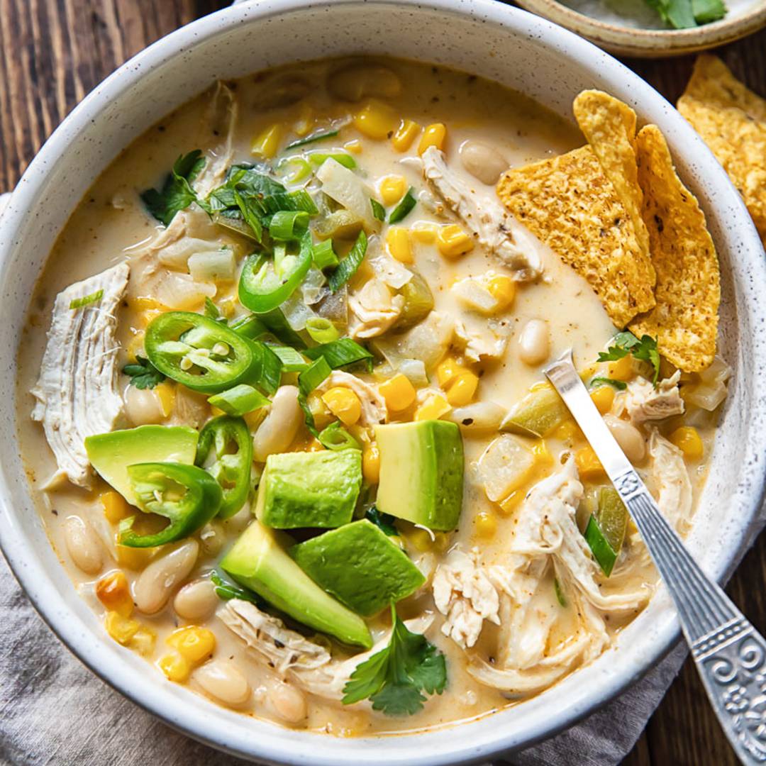 top down view of a bowl of white chili with tortillas, avocados and chopped peppers