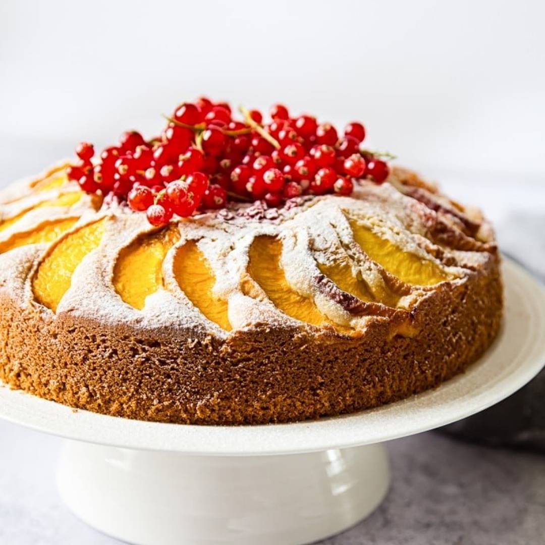 A cake studded with peaches and topped with red berries and a sugar dusting on a cake stand.