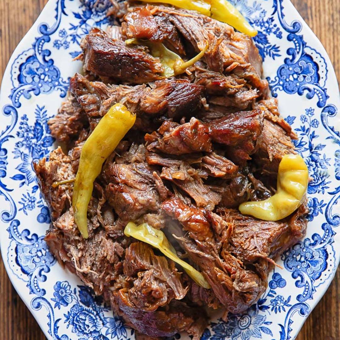 Blue and white platter filled with slow cooked beef and long yellow peppers on a wooden background.