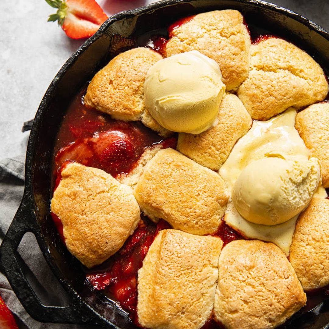 A skillet holding a cobbler made with red strawberries, topped with doughy biscuits and iced cream.