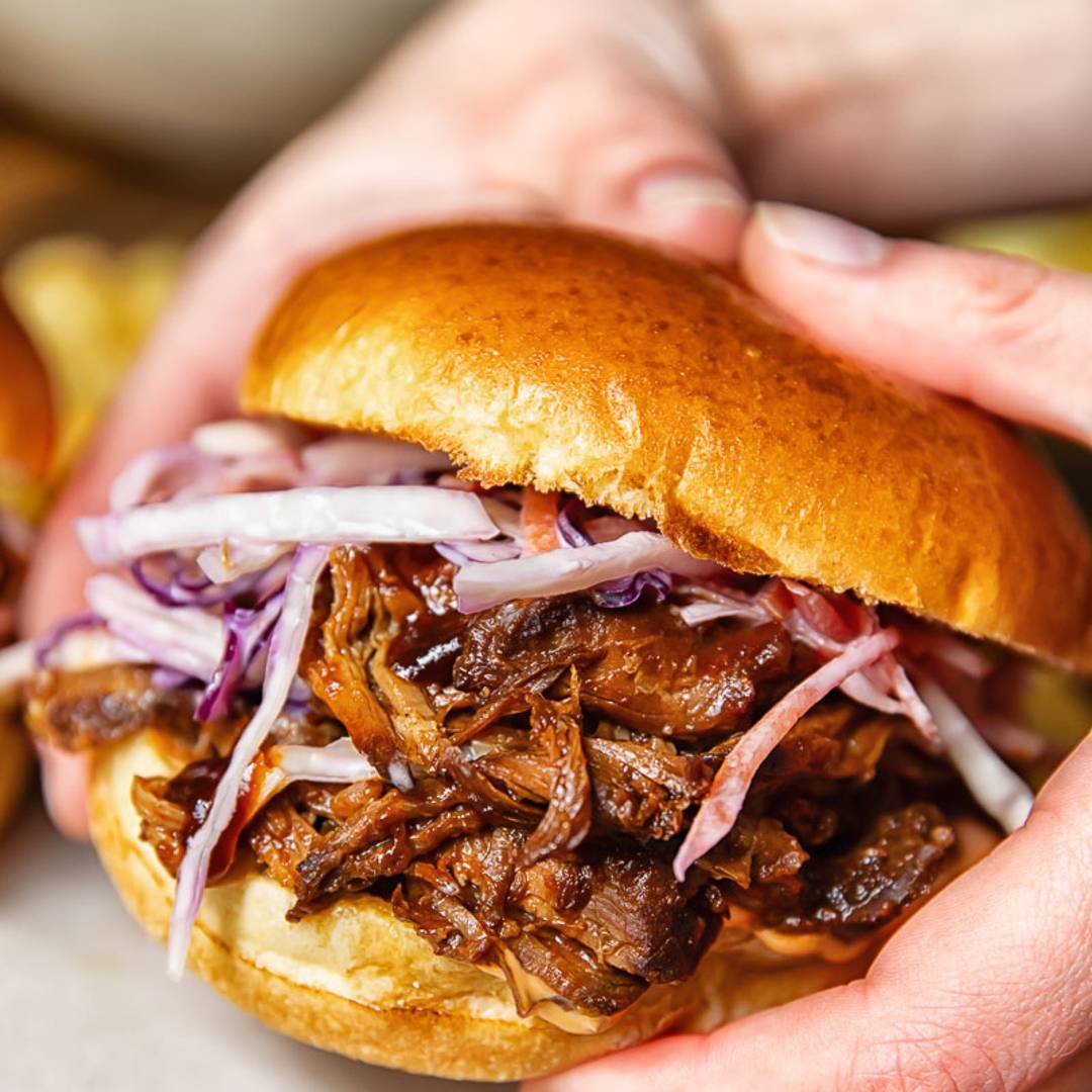 hands holding a sandwich made with shredded pork and slaw between a brioche bun.