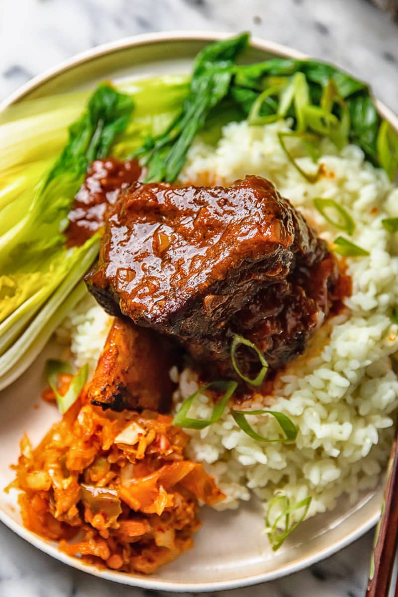A short rib covered in sauce on a bed of rice, kimchi and greens.