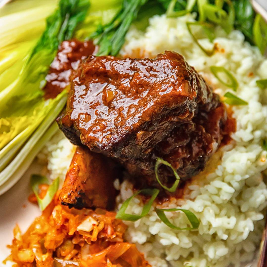 A short rib covered in sauce on a bed of rice, kimchi and greens.