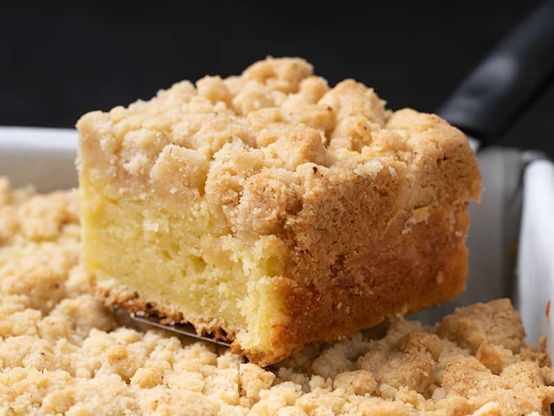 Square of yellow cake with light brown crumble topping on spatula with rest of cake in white paper below
