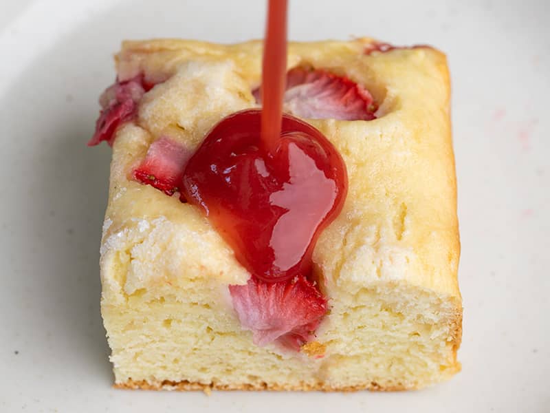 strawberry syrup pouring on square of strawberry breakfast cake on white plate
