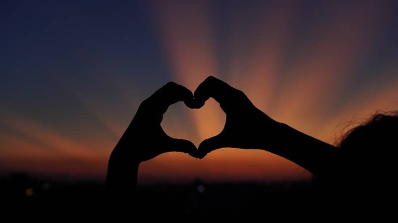 Hands form a heart shape against the sunset.